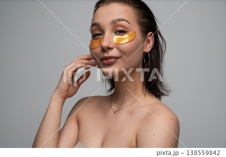 Young woman applying gold eye patches, caring for her skin and promoting a youthful look while enjoying a beauty routine for self-care and wellness against a white background Young woman applying gold eye patches, caring for her skin and promoting a youthful look while enjoying a beauty routine for self-care and wellness against a white background 138559842