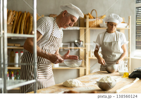 Two bakers prepare dough for baguettes - whisking eggs and sprinkling flour over the dough through sieve 138560124