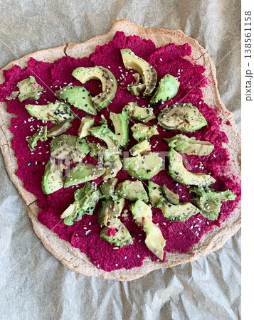 A round pie base features beet hummus spread evenly on top. Fresh avocado slices are arranged on the hummus. This dish is served on a paper-lined surface during midday meal preparation A round pie base features beet hummus spread evenly on top. Fresh avocado slices are arranged on the hummus. This dish is served on a paper-lined surface during midday meal preparation 138561158