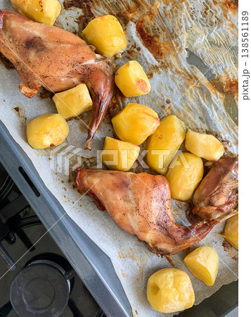 Rabbit pieces with potatoes are placed on a baking tray lined with parchment paper in an oven. The meat is seasoned with herbs like thyme for flavor 138561189