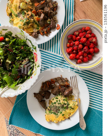 A meal features beef liver with a zucchini omelette on a white plate, fresh salad in a bowl, and strawberries for dessert on a table with a blue and white tablecloth A meal features beef liver with a zucchini omelette on a white plate, fresh salad in a bowl, and strawberries for dessert on a table with a blue and white tablecloth 138561196