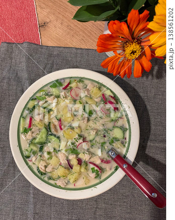 A bowl filled with okroshka, a cold Russian soup, shows fresh ingredients like vegetables and herbs. It is placed on a table with a touch of flowers adding some color A bowl filled with okroshka, a cold Russian soup, shows fresh ingredients like vegetables and herbs. It is placed on a table with a touch of flowers adding some color 138561202