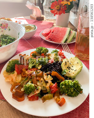 A plate features brown rice topped with baked vegetables and avocado. Watermelon and a drink sit nearby. The table includes bowls of broccoli and a bright flower arrangement A plate features brown rice topped with baked vegetables and avocado. Watermelon and a drink sit nearby. The table includes bowls of broccoli and a bright flower arrangement 138561203