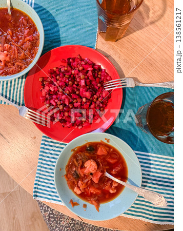 A bowl of Ukrainian borscht sits next to a plate of colorful salad and another bowl of food on a wooden table. Glasses filled with a beverage are placed around the dishes 138561217