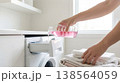 Person pours pink liquid detergent into laundry machine drawer while folded towels are stacked on countertop. Bright room with natural light coming from window. 138564059