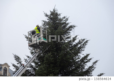 Professional worker operates hydraulic lift, carefully decorating grand outdoor evergreen tree with sparkling Christmas lights for festive holiday season celebration Professional worker operates hydraulic lift, carefully decorating grand outdoor evergreen tree with sparkling Christmas lights for festive holiday season celebration 138564614