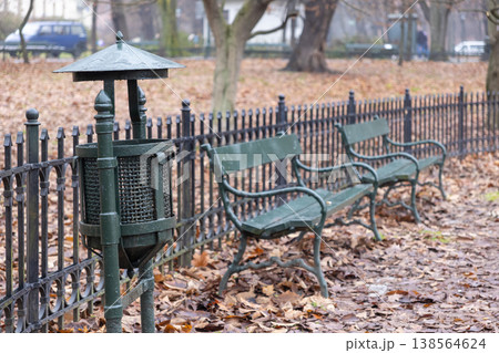 Dark green metal trash bin and two classic park benches beside a wrought-iron fence, covered in fallen autumn leaves in a peaceful urban park Dark green metal trash bin and two classic park benches beside a wrought-iron fence, covered in fallen autumn leaves in a peaceful urban park 138564624
