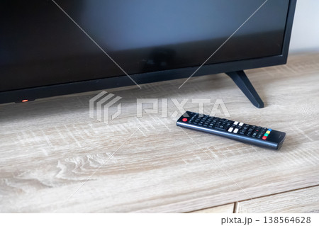 Black television screen with a red indicator light on standby and a modern remote control resting on a light wooden table, representing home leisure Black television screen with a red indicator light on standby and a modern remote control resting on a light wooden table, representing home leisure 138564628