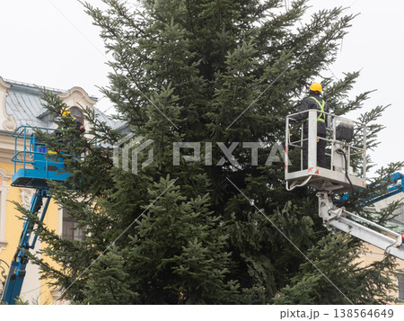 Expert team members on mobile elevated work platforms meticulously adorn a giant evergreen tree with shimmering festive lights, preparing public spaces for upcoming celebrations Expert team members on mobile elevated work platforms meticulously adorn a giant evergreen tree with shimmering festive lights, preparing public spaces for upcoming celebrations 138564649