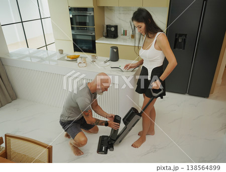 Man crouching cleaning vacuum while woman watches in bright kitchen Man crouching cleaning vacuum while woman watches in bright kitchen 138564899