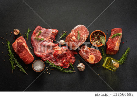 Different cuts of beef steak are arranged on a black surface. Fresh herbs, spices, and garlic accompany the meat, ready for cooking or grilling at a kitchen or market Different cuts of beef steak are arranged on a black surface. Fresh herbs, spices, and garlic accompany the meat, ready for cooking or grilling at a kitchen or market 138566685