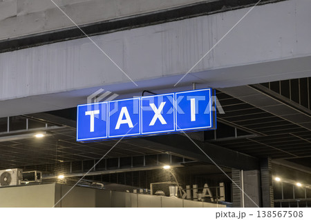 Bright blue illuminated taxi sign with white letters hanging from an urban structure, indicating transportation services and convenient traveler pick-up guidance 138567508