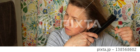 A 50-year-old woman styles her hair with a straightener in a bathroom. Floral curtains add color to the space. She is focused on her hair, preparing for the day ahead, banner A 50-year-old woman styles her hair with a straightener in a bathroom. Floral curtains add color to the space. She is focused on her hair, preparing for the day ahead, banner 138568602