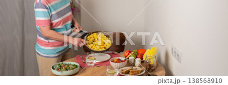 A 53-year-old woman prepares dinner at a round wooden table in her kitchen. She pours food from a colander onto a plate while arranging utensils and other items, banner A 53-year-old woman prepares dinner at a round wooden table in her kitchen. She pours food from a colander onto a plate while arranging utensils and other items, banner 138568910