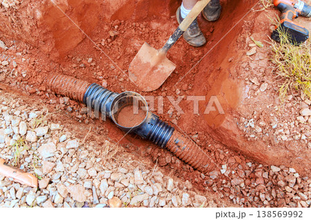 Worker digs trench for drainage pipe installation in red dirt, section of black pipe, tools nearby. Worker digs trench for drainage pipe installation in red dirt, section of black pipe, tools nearby. 138569992