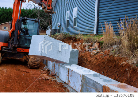 Construction workers use excavator to lift place concrete blocks for retaining walls beside house. Construction workers use excavator to lift place concrete blocks for retaining walls beside house. 138570109