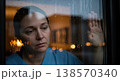 Sad woman standing by a window and watching rain outside. Thoughtful mood and isolation. Concept of burnout, stress, and mental health. 138570340