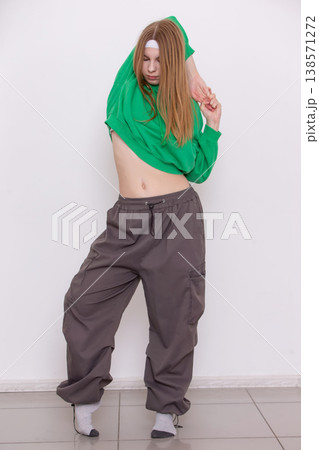 Girl in green long-sleeve top and gray baggy pants strikes a stylish dance pose indoors Girl in green long-sleeve top and gray baggy pants strikes a stylish dance pose indoors 138571272