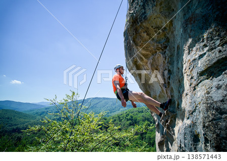 Aerial view of male rock climber ascending rugged limestone cliff with harness and rope for safety. Sportsman climbing on vertical large boulder at Dobvush Rocks in Carpathian mountains, Ukraine. Aerial view of male rock climber ascending rugged limestone cliff with harness and rope for safety. Sportsman climbing on vertical large boulder at Dobvush Rocks in Carpathian mountains, Ukraine. 138571443