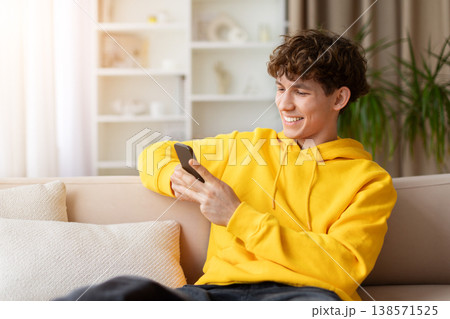 Happy teen boy in yellow hoodie sitting on sofa using smartphone 138571525
