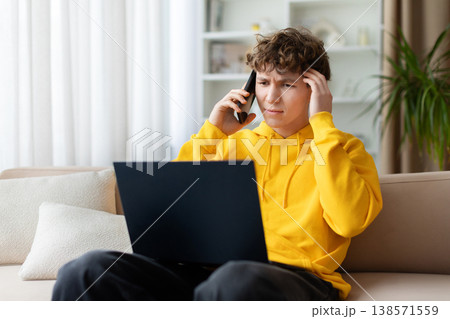 Stressed teen boy in yellow hoodie talking on phone with laptop 138571559