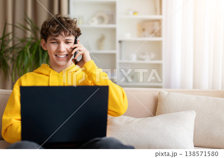 Happy teen boy talking on phone while using laptop at home 138571580