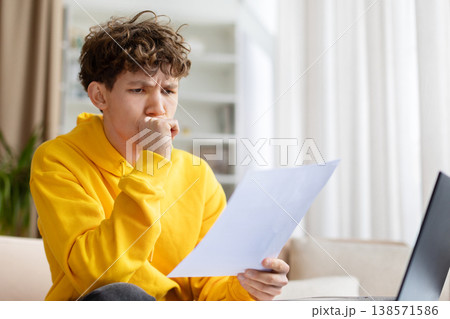 Anxious teen boy reading paper document at laptop with hand on mouth Anxious teen boy reading paper document at laptop with hand on mouth 138571586