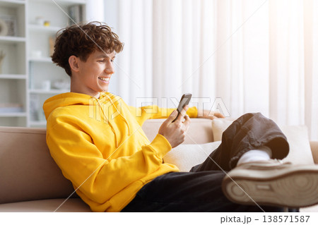 Smiling young teen male in yellow hoodie sitting on couch using smartphone 138571587