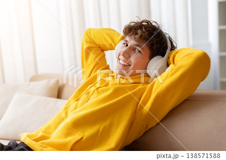 Relaxed teen boy wearing wireless headphones and hoodie leaning back on sofa 138571588