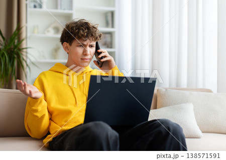 Confused frustrated teen boy in yellow hoodie on phone call with laptop 138571591