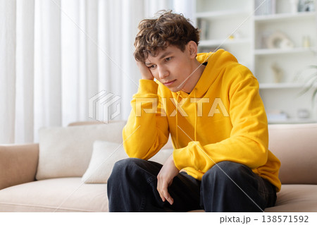 Sad bored teen boy sitting alone on sofa with hand on cheek Sad bored teen boy sitting alone on sofa with hand on cheek 138571592