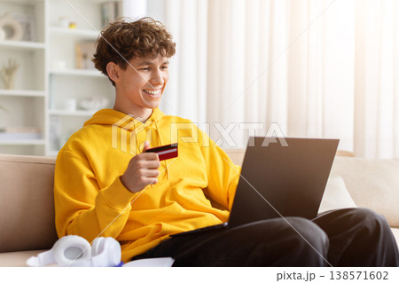 Happy young teen guy holding credit card at laptop on sofa, smiling 138571602