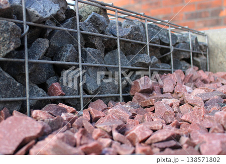 Small gabion fence from steel mesh filled with gray granite stones on a smooth surface of red granite rocks on a floor. Ornate Stones for Outdoor Landscape Design Small gabion fence from steel mesh filled with gray granite stones on a smooth surface of red granite rocks on a floor. Ornate Stones for Outdoor Landscape Design 138571802