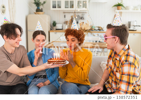 Make a wish. Woman wearing party cap blowing out burning candles on birthday cake. Happy Birthday party. Group of friends wishes girl happy birthday. People celebrating birthday with party at home Make a wish. Woman wearing party cap blowing out burning candles on birthday cake. Happy Birthday party. Group of friends wishes girl happy birthday. People celebrating birthday with party at home 138572351