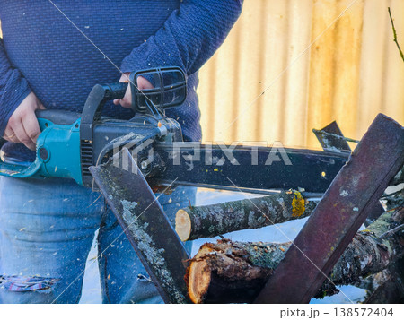 Man actively using an electric chainsaw to cut tree branches and logs into smaller pieces for firewood, working outdoors with sawdust flying and preparing wood for a home heating project. 138572404