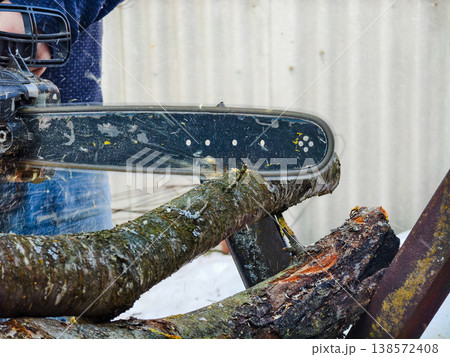 Person operating a powerful chainsaw, cutting through a thick tree branch for firewood during cold winter weather, demonstrating manual labor and preparation for heating. Person operating a powerful chainsaw, cutting through a thick tree branch for firewood during cold winter weather, demonstrating manual labor and preparation for heating. 138572408