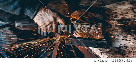 Industrial worker wearing safety gloves operating an angle grinder, producing a shower of bright orange sparks while shaping a metal profile in a workshop. Industrial worker wearing safety gloves operating an angle grinder, producing a shower of bright orange sparks while shaping a metal profile in a workshop. 138572413