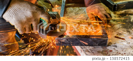 Skilled laborer wearing protective gloves operating an angle grinder to cut or finish metal material, creating a bright shower of sparks in an industrial workshop environment. Skilled laborer wearing protective gloves operating an angle grinder to cut or finish metal material, creating a bright shower of sparks in an industrial workshop environment. 138572415