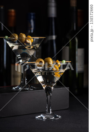 Two martini glasses with skewered green olives and citrus twists against dark surface with wine bottles blurred in the background. 138572660