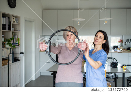 Senior woman performing physiotherapy exercise with ring under physiotherapist supervision at home. 138572740