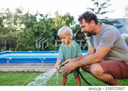 Father and toddler son watering garden with hose near backyard pool. 138572867