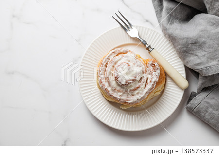 Freshly baked cinnamon roll with white icing on a plate, fork and napkin on a marble background. 138573337