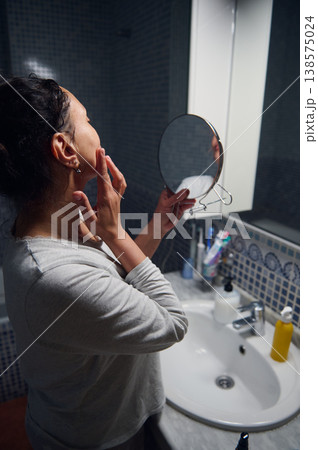 Middle-Aged Woman in Pyjamas Checking Her Face in Bathroom Mirror During Morning Beauty Routine 138575024