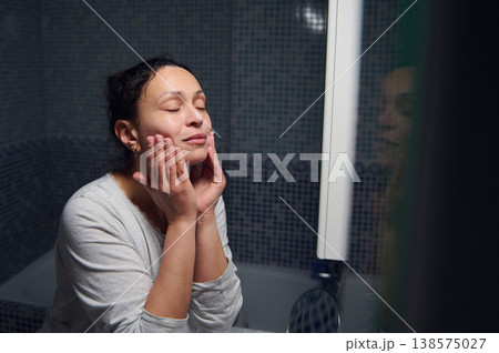 Middle-Aged Woman in Pajamas Doing Morning Skincare Routine at Bathroom Mirror 138575027