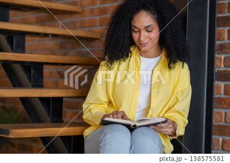 Pretty brunette young woman sitting on the wooden stairs with a book Pretty brunette young woman sitting on the wooden stairs with a book 138575581