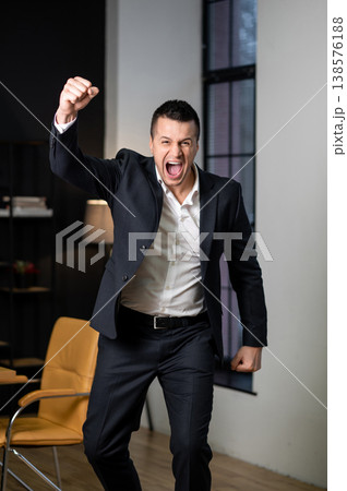 Excited businessman standing with raised arms celebrating success in office interior 138576188