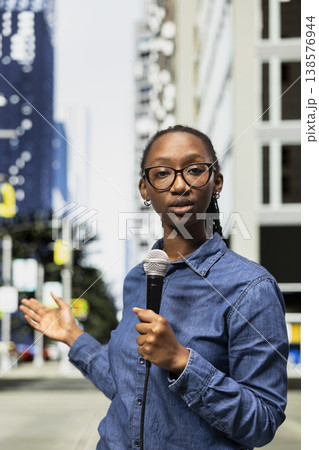 Field reporter holds mic, speaking towards camera on city street, presenting information. African american journalist reports live news update with microphone from metropolitan downtown area avenue Field reporter holds mic, speaking towards camera on city street, presenting information. African american journalist reports live news update with microphone from metropolitan downtown area avenue 138576944