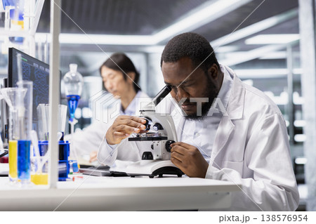 Materials scientist using microscope to evaluate cellular structures in lab workspace. African american man observes specimens through magnification tool at laboratory bench workstation Materials scientist using microscope to evaluate cellular structures in lab workspace. African american man observes specimens through magnification tool at laboratory bench workstation 138576954
