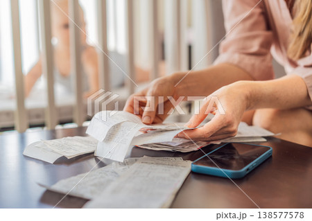 Mother sitting on sofa calculating monthly expenses with receipts while baby lies in crib nearby at home. Stressed mom managing budget and childcare. Financial pressure and parenthood concept Mother sitting on sofa calculating monthly expenses with receipts while baby lies in crib nearby at home. Stressed mom managing budget and childcare. Financial pressure and parenthood concept 138577578