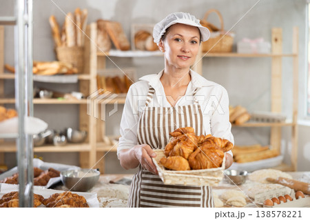 Portrait of young happy female baker working in bakehouse, holding tray with fresh bakery goods on kitchen 138578221